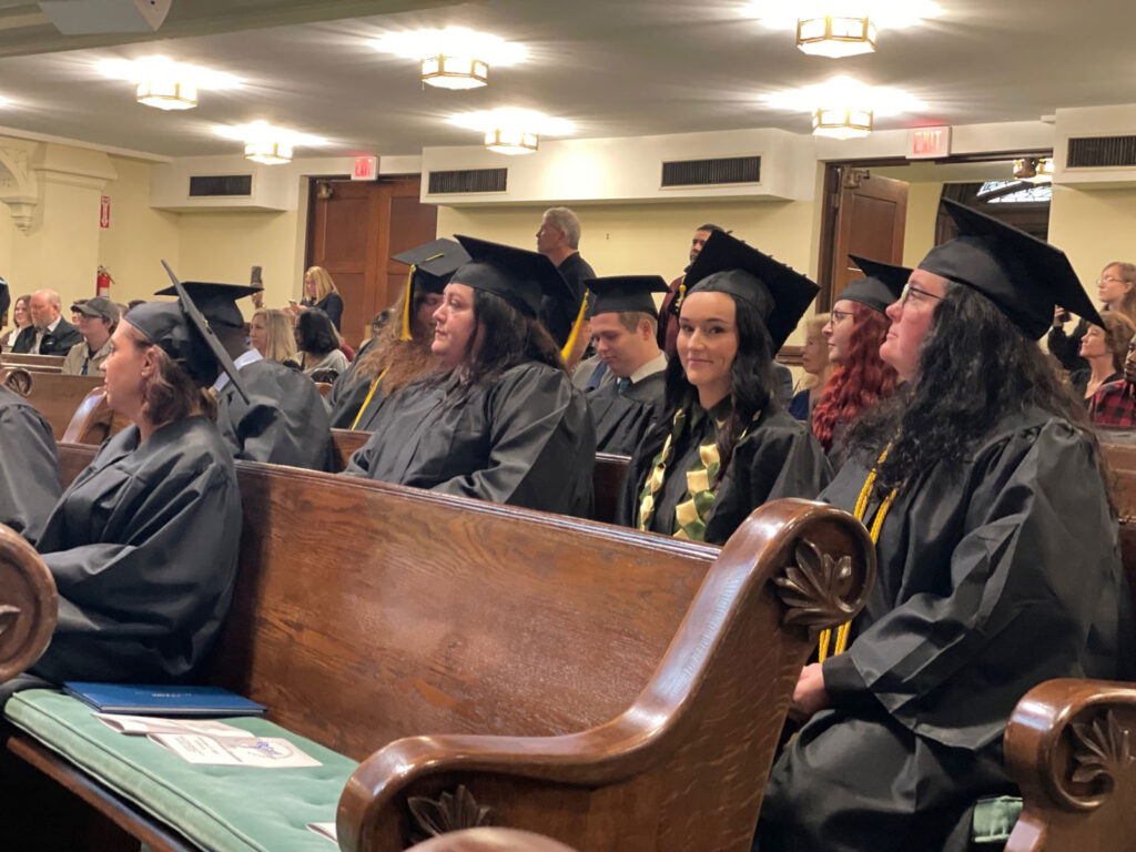 Graduating students wearing black gowns and caps, sitting in benches waiting for the ceremony to being.